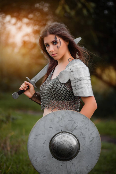  Armed medieval woman warrior in armor posing while standing on the background of bokeh of the forest and sunset.  - Foto, afbeelding