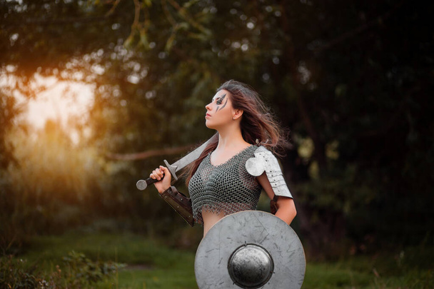  Armed medieval woman warrior in armor posing while standing on the background of bokeh of the forest and sunset.  - Φωτογραφία, εικόνα