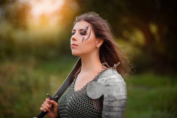  Armed medieval woman warrior in armor posing while standing on the background of bokeh of the forest and sunset.  - Foto, imagen