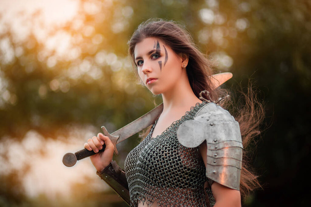  Armed medieval woman warrior in armor posing while standing on the background of bokeh of the forest and sunset.  - Fotoğraf, Görsel