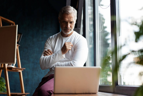 Alkalmi ősz hajú, szemüveges, idős férfi, aki laptopon dolgozik, miközben az ablak melletti irodában vagy kávézóban ül. - Fotó, kép