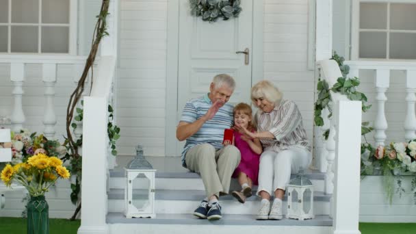 Pareja mayor sentada con su nieta en el porche de su casa. Uso del teléfono móvil para videollamadas en línea - Imágenes, Vídeo