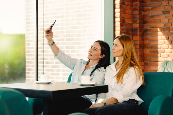 Excited friends make selfie on smartphone having fun in coffeeshop. Students smile for picture on phone met together in cafe. Two young business women posing for self-portrait in office coffee break.  - 写真・画像