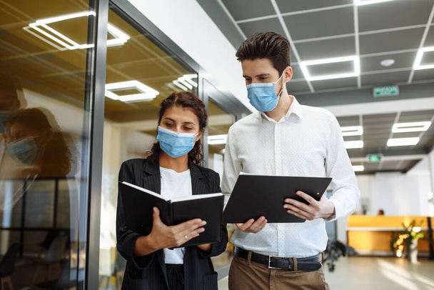 企業家は、コロナウイルスのパンデミックの間の労働問題について議論する。男と女は防護医療マスクを着てオフィスの廊下でビジネスを話している。職場の安全衛生コンセプト - 写真・画像