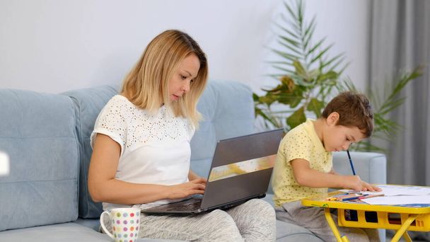 Smiling mom working at home with her child on the sofa while writing an email. Young woman working from home, while in quarantine isolation. Calm young mother or nanny sit on couch working on laptop - Photo, Image