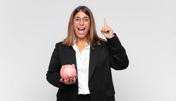 young woman with a piggy bank feeling like a happy and excited genius after realizing an idea, cheerfully raising finger, eureka! - Photo, image
