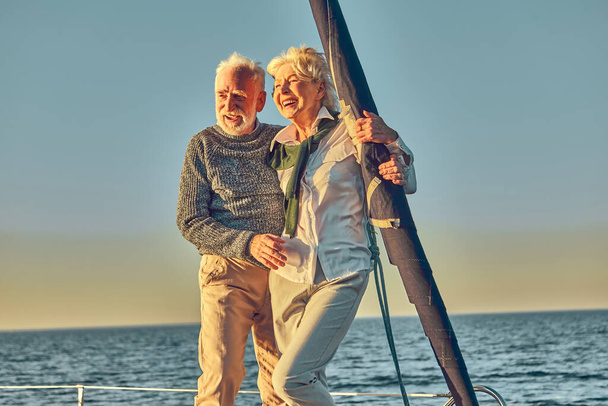 Happy senior couple in love, retired man and woman standing on the side of sail boat or yacht deck floating in sea, enjoying amazing sunset and smiling - Photo, Image