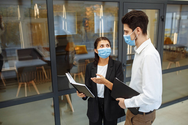 Двое коллег обсуждают работу и бизнес в защитных стерильных медицинских масках, стоящих в коридоре офиса. Коронавирус предотвращает распространение во время глобальной пандемии - Фото, изображение