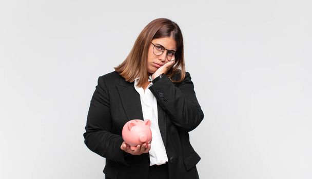 young woman with a piggy bank feeling bored, frustrated and sleepy after a tiresome, dull and tedious task, holding face with hand - Фото, зображення