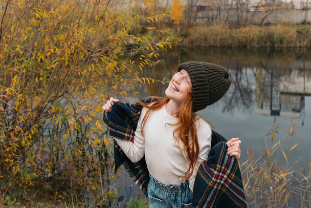 Happy red-haired girl in a knitted hat on an autumn walk by the lake. - Photo, Image