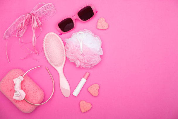 Flat lay of pink female bathroom accessories lying on a pink field. Accessories of every woman including lipstick, hairbrush, sunglasses, sponge, soap, and headband - Photo, Image