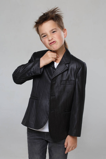 Little stylish smiling boy in a jacket grimaces and grabs his neck with his hand isolated on a gray background. - 写真・画像