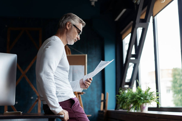Casual Grey-haired Mature man reading paper in his office. - 写真・画像