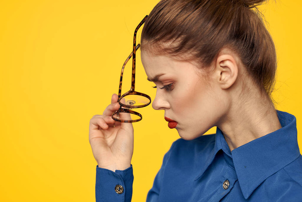 Ritratto di donna con gli occhiali tra le mani su sfondo giallo e in camicia blu vista ritagliata - Foto, immagini