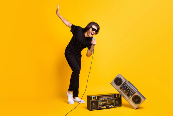 Full length photo of young man hold mic open mouth raise hand two retro radio wear black t-shirt pants white sneakers glasses isolated yellow color background - Фото, зображення