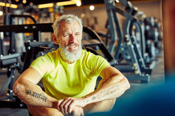 Portret mężczyzny w średnim wieku omawiającego plan treningu z instruktorem fitness podczas siedzenia razem na podłodze na siłowni - Zdjęcie, obraz