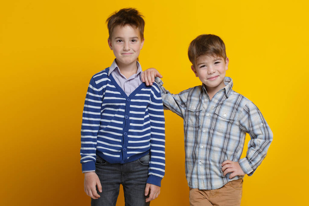 Two funny little cheerful boys are standing side by side, one is leaning on the shoulder of the second, isolated on a bright yellow background. - Foto, immagini