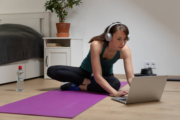 Mulheres bonitas fazendo fitness em casa perto da cama e assistindo tutoriais on-line no laptop em luz natural. Esporte e conceito de recreação. - Foto, Imagem
