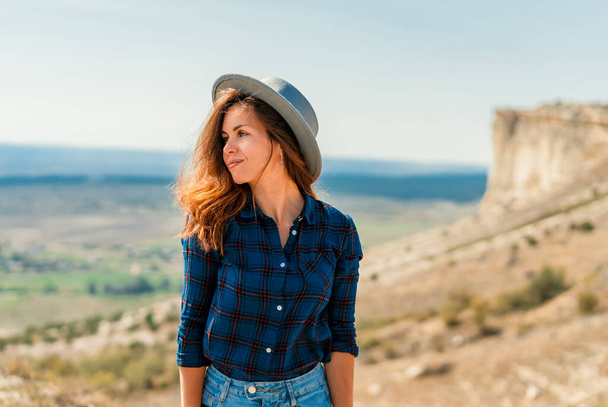 A young woman in a plaid shirt and hat stands on a mountain with a view of the rustic landscape, White rock in the Crimea - Foto, Bild