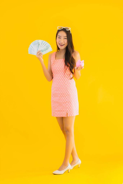 Portret piękna młoda azjatycka kobieta pokazać dużo gotówki lub pieniędzy z świnki bank na żółtym odizolowanym tle - Zdjęcie, obraz