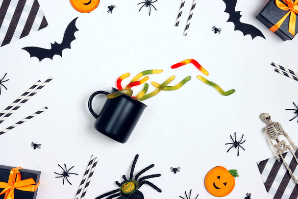 Black mug with gummy worm surrounded by Halloween decorations on a white table. Halloween party food and drink. - Fotó, kép
