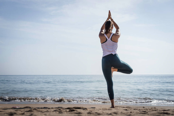 Giovane donna sportiva che fa esercizi di Yoga utilizzando un tappetino da palestra lungo la spiaggia di Lisbona, Portogallo. Donna giocosa che lavora come insegnante di yoga freelance facendo allenamento di fitness sulla spiaggia al tramonto. Vita sana - Foto, immagini