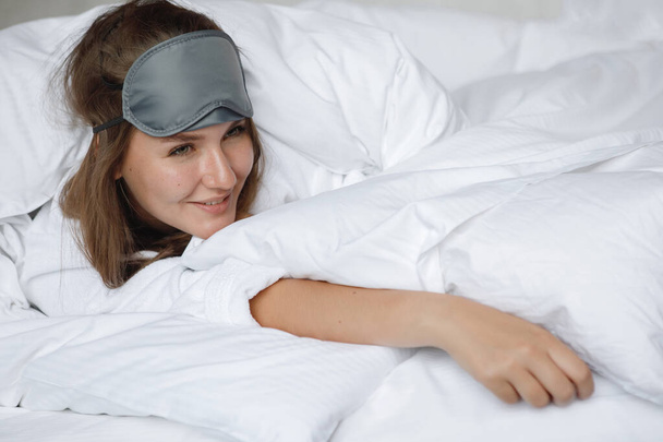 Γυναίκα με λευκή ρόμπα και μάσκα ύπνου ξύπνησε και χαμογέλασε στο κρεβάτι το πρωί σε ένα δωμάτιο ξενοδοχείου - Φωτογραφία, εικόνα