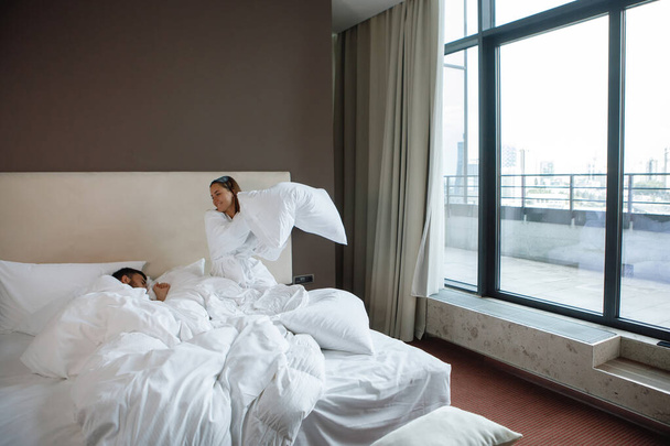 Γυναίκα και ένας άνδρας σε ένα λευκό κρεβάτι παίζουν με ένα μαξιλάρι κοντά σε ένα μεγάλο παράθυρο. Υπόλοιπο στο ξενοδοχείο, το ζευγάρωμα παιχνίδια. - Φωτογραφία, εικόνα