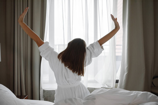 Жінка в білому халаті, що розтягується, прокидається на ліжку перед вікном. Вид ззаду
. - Фото, зображення