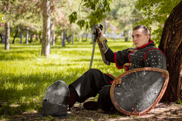 A medieval warrior with a saber and a shield in his hands sits in the shadows on the grass against the background of the forest. The eastern knight is resting with his back against a tree. - Фото, зображення