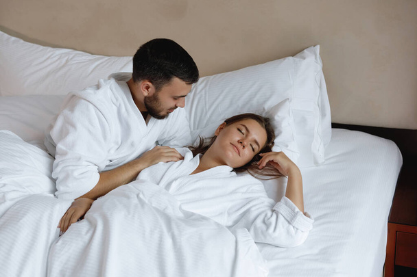 Ζευγάρι στο κρεβάτι. Νεαρός γενειοφόρος άντρας και γυναίκα ξαπλώνουν σε λευκό μαξιλάρι και ξυπνάνε νωρίς το πρωί.. - Φωτογραφία, εικόνα