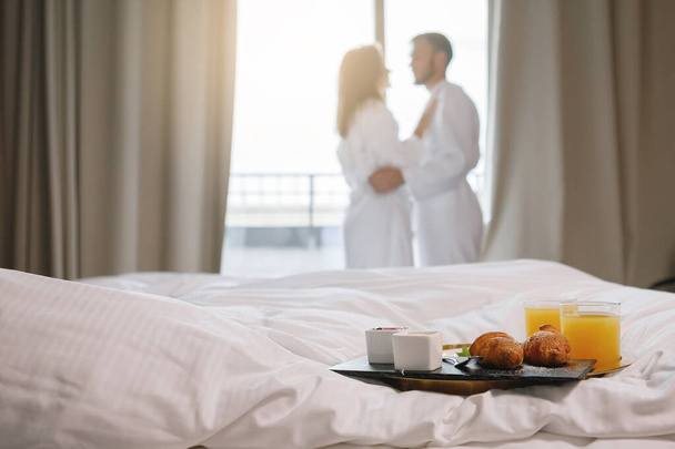 Breakfast tray is on a white bed. A couple in bathrobes are drinking coffee by the window in the background. Selective focus. - Foto, Bild