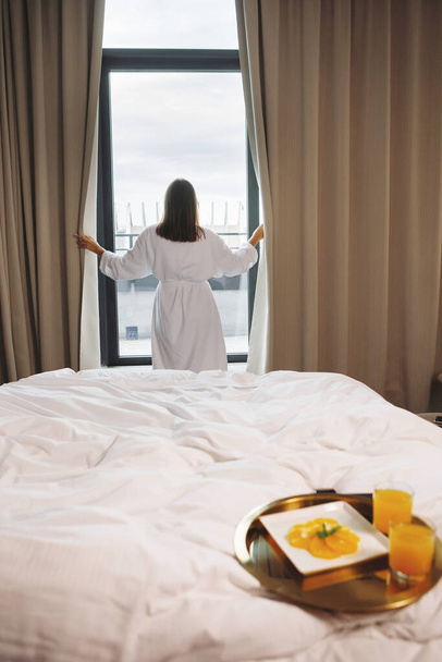 Taca śniadaniowa jest na białym łóżku. Kobieta w szlafroku rozsuwa zasłony przy oknie w tle. Skupienie selektywne. - Zdjęcie, obraz