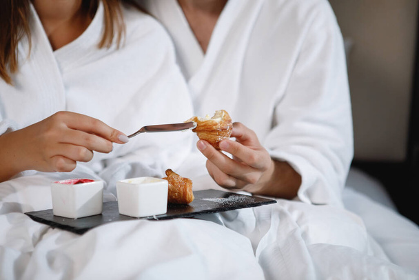 Bearded man and a woman in white coats are having breakfast in bed. Selective focus. Clouse-up. - Valokuva, kuva