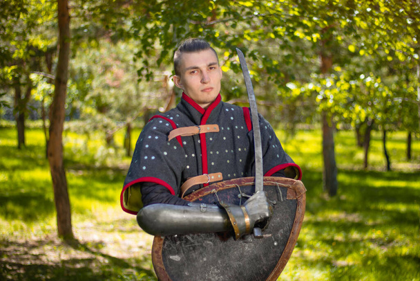 Portrait of a medieval nomad warrior in armor with a shield and saber in his hands against the background of the forest. - Foto, afbeelding