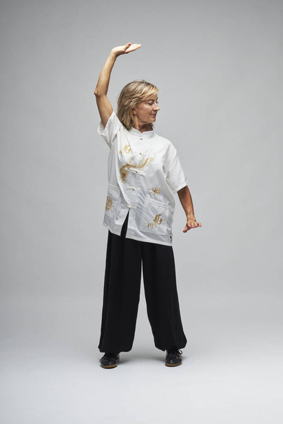 Mature blonde woman practicing Chi kung and Tai Chi on a white background. She wears a traditional white chinese Tai Chi jacket, black trousers and black shoes with ying yang symbol - Foto, afbeelding