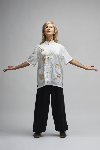 Mature blonde woman practicing Chi kung and Tai Chi on a white background. She wears a traditional white chinese Tai Chi jacket, black trousers and black shoes with ying yang symbol - 写真・画像