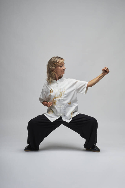 Mature blonde woman practicing Chi kung and Tai Chi on a white background. She wears a traditional white chinese Tai Chi jacket, black trousers and black shoes with ying yang symbol - Foto, Imagem