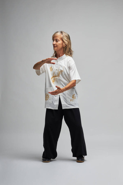 Ältere blonde Frau praktiziert Chi Kung und Tai Chi auf weißem Hintergrund. Sie trägt eine traditionelle weiße chinesische Tai-Chi-Jacke, schwarze Hose und schwarze Schuhe mit Ying-Yang-Symbol - Foto, Bild