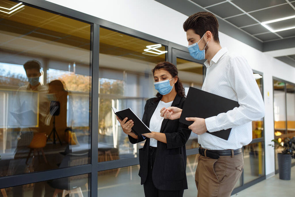Бизнесмены обсуждают рабочие вопросы во время пандемии коронавируса. Мужчина и женщина в защитных медицинских масках говорят о делах в офисном коридоре. Безопасность труда на производстве - Фото, изображение