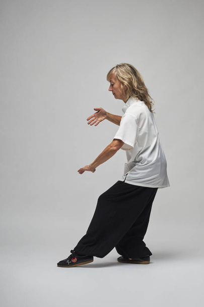 Starší blondýny žena cvičí Chi kung a Tai Chi na bílém pozadí. Nosí tradiční bílou čínskou bundu Tai Chi, černé kalhoty a černé boty se symbolem ying Yang - Fotografie, Obrázek