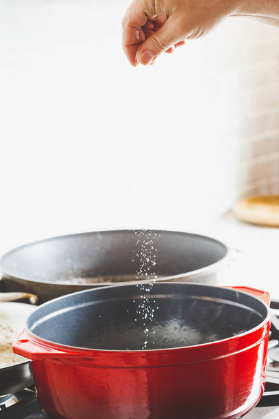 Main d'homme ajoutant du sel lors de la cuisson des aliments dans un four français en fonte émaillée rouge - Photo, image