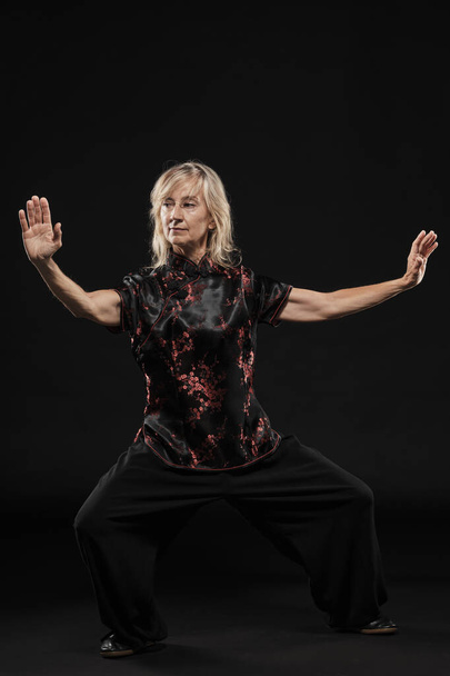 Mature blonde woman practicing Tai Chin Chen style in a black background wearing a traticional chinese black jacket with red decoration, black trousers and traditional shoes with ying yang symbol - Photo, Image