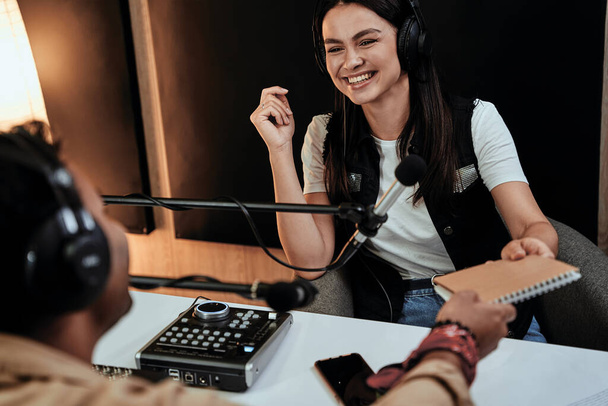 Portret van een vrolijke vrouwelijke radio-presentator die lacht terwijl hij praat met mannelijke gast, presentator en een notitieboekje aan hem geeft, terwijl hij een live show modereert in de studio - Foto, afbeelding