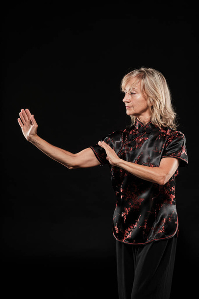 Mature blonde woman practicing Tai Chin Chen style in a black background wearing a traticional chinese black jacket with red decoration, black trousers and traditional shoes with ying yang symbol - Photo, Image