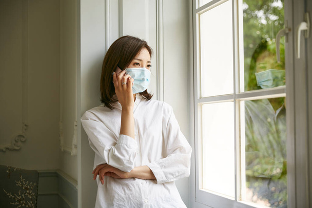 mladá asijská žena v izolaci nosí masku stojící u okna mluví na mobilu - Fotografie, Obrázek