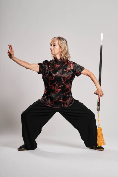Mature blonde woman practicing Tai Chin with sword in a white background wearing a traditional chinese black jacket with red chinese decoration, black trousers and chinese shoes with ying yang symbol - Valokuva, kuva