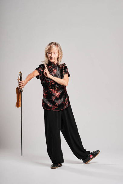 Mature blonde woman practicing Tai Chin with sword in a white background wearing a traditional chinese black jacket with red chinese decoration, black trousers and chinese shoes with ying yang symbol - Photo, Image