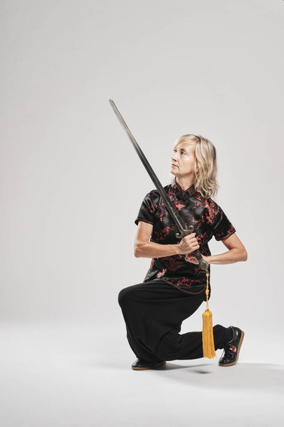 Mature blonde woman practicing Tai Chin with sword in a white background wearing a traditional chinese black jacket with red chinese decoration, black trousers and chinese shoes with ying yang symbol - Фото, зображення