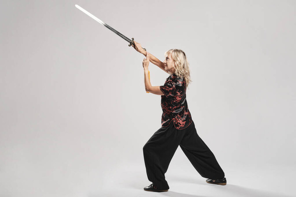 Mature blonde woman practicing Tai Chin with sword in a white background wearing a traditional chinese black jacket with red chinese decoration, black trousers and chinese shoes with ying yang symbol - Fotó, kép